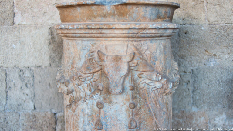 Cylindrical carved stone pedestal with a bull’s skull and hanging garland relief against a masonry wall.