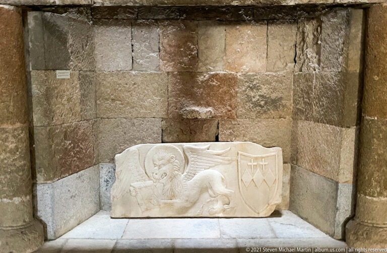 Rectangular stone relief with a reclining lion beside a shield bearing a cross-like heraldic device.
