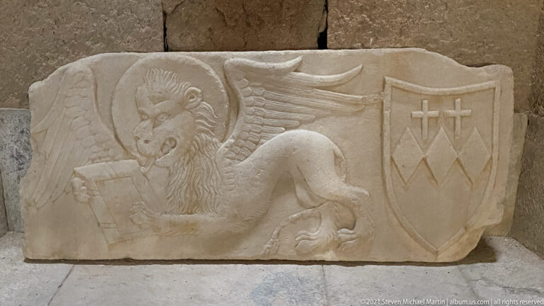 Stone relief of a haloed winged lion holding an open book beside a shield with a cross and chevron-like device.
