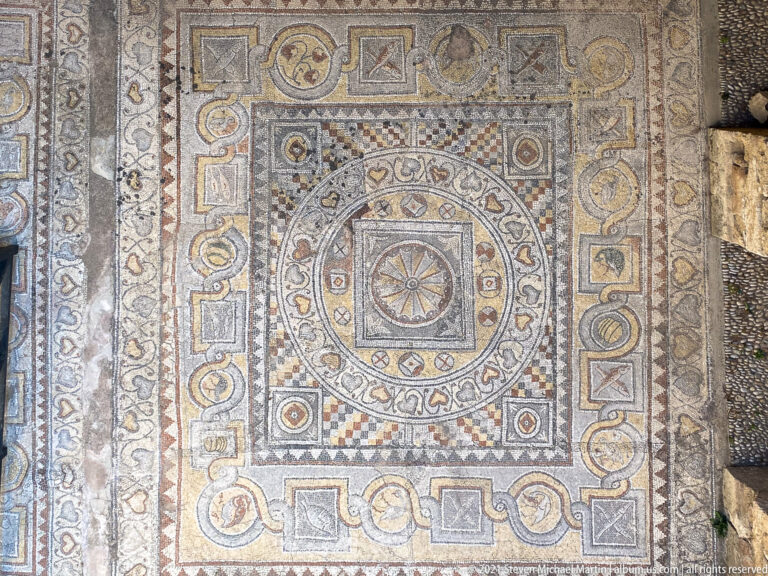 Large geometric mosaic pavement with central rosette, concentric circles, checkerboard fields, braided borders, and small medallions.