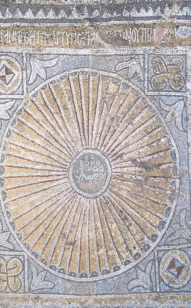 Circular mosaic with many golden rays around a central Greek inscription, bordered by scallops and surrounding geometric panels.