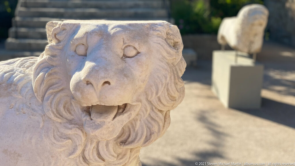 Close frontal view of a marble lion’s face with deeply carved mane, rounded eyes, open mouth, and visible teeth.