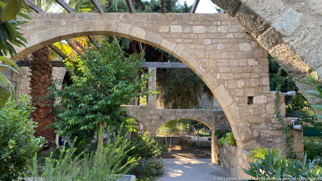 Large stone arch over a lush garden path with a smaller arch beyond, surrounded by palms and dense greenery.