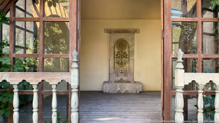 Ornamental vertical stone panel with circular rosette and floral carving displayed inside an open wooden garden pavilion.