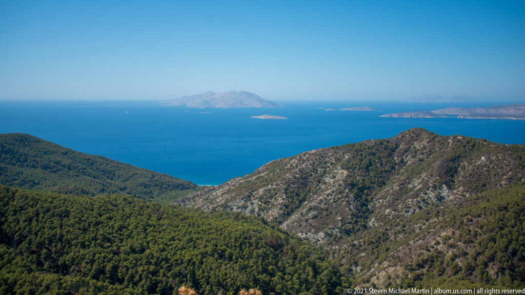 Pine-covered limestone ridges descend toward the blue Aegean, with several low islands visible on the horizon.