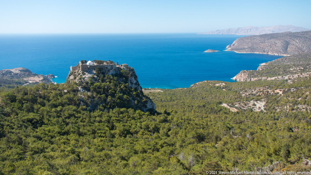 Small stone castle atop a wooded crag, surrounded by pine forest with blue sea, headlands, and an offshore islet beyond.