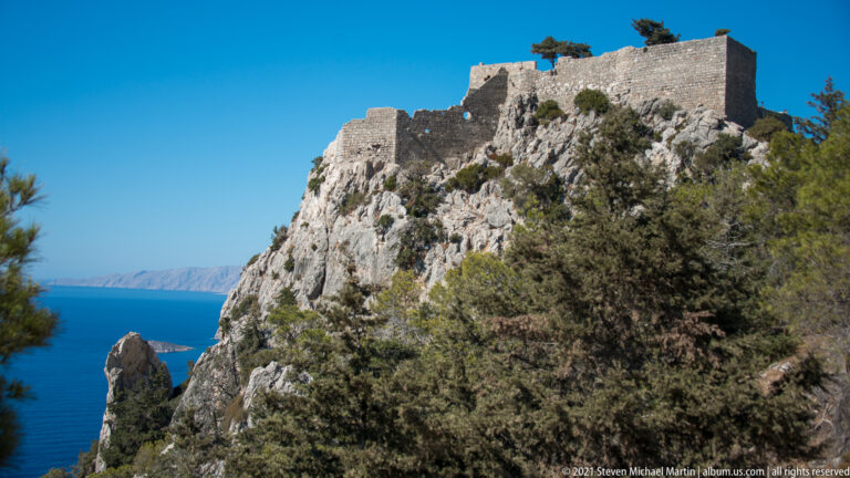 Stone walls of Monolithos Castle rise above a steep limestone cliff and trees, with a glimpse of blue sea at left.