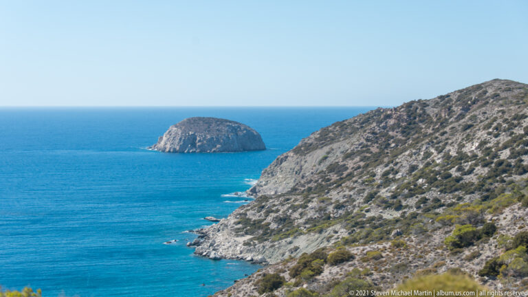 Rounded rocky islet offshore from a steep scrub-covered limestone headland beside clear blue water.