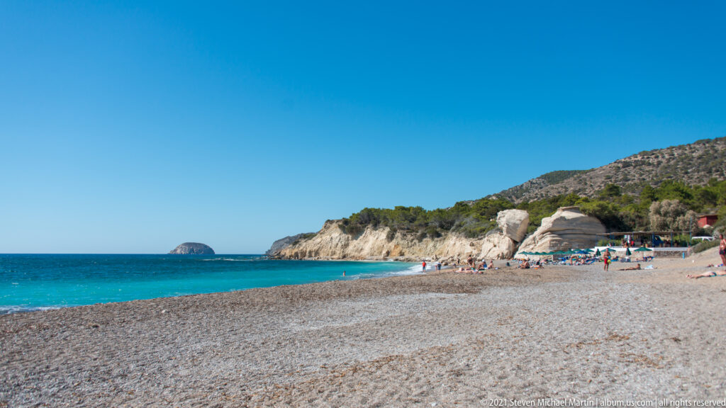 Wide pebble beach with swimmers, umbrellas, pale cliffs, turquoise sea, and a rounded islet offshore.
