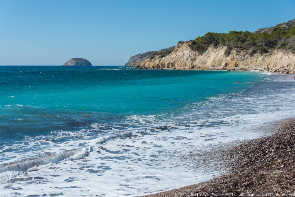 Turquoise waves wash onto a pebble beach below pale cliffs, with a rounded rocky islet visible offshore.