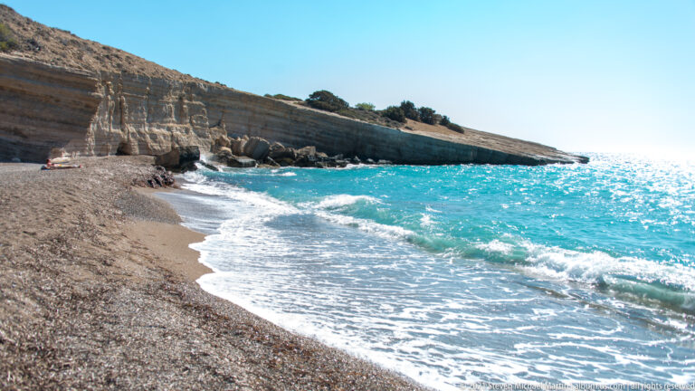 Layered pale rock cliff projects beside a pebble beach as turquoise waves break around fallen blocks at its base.