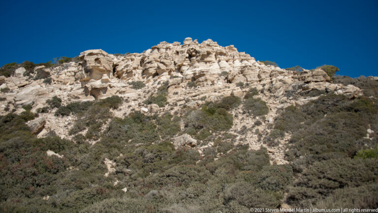 Pale weathered limestone outcrop with irregular cavities and ledges rising above dense low Mediterranean scrub.
