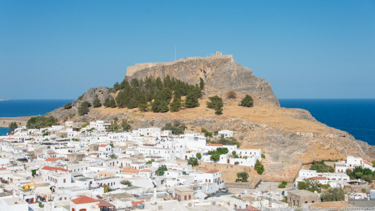 Whitewashed Lindos village below a rocky acropolis crowned by massive stone fortifications and columnar ruins.