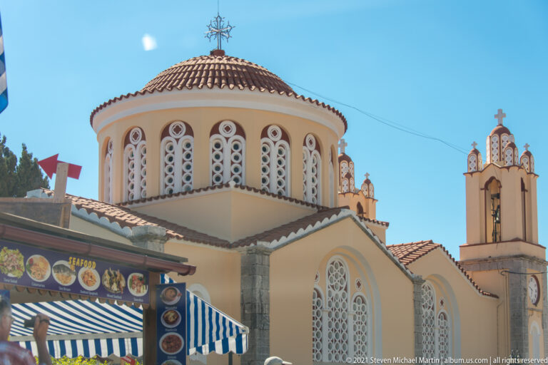 Cream-colored Orthodox church with a tiled octagonal dome, patterned arched windows, and a narrow bell tower above a shopfront.