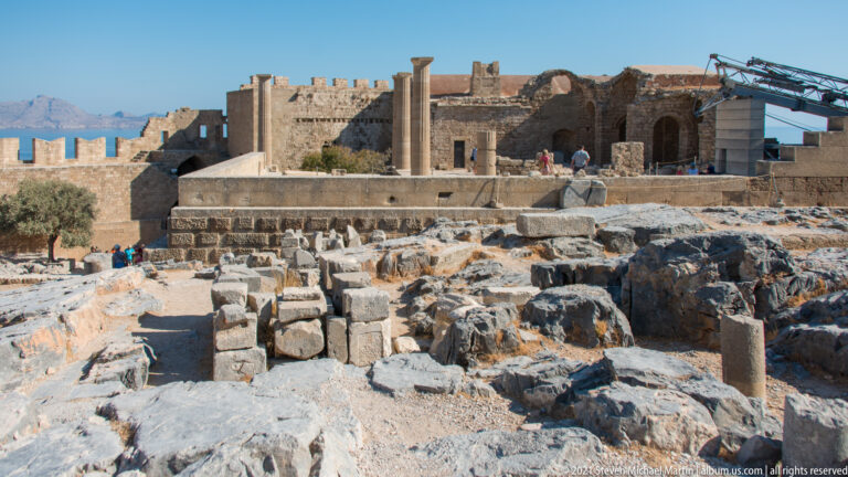 Standing stone columns, foundations, rock-cut surfaces, and fallen blocks inside the fortified Acropolis of Lindos, with visitors beyond.