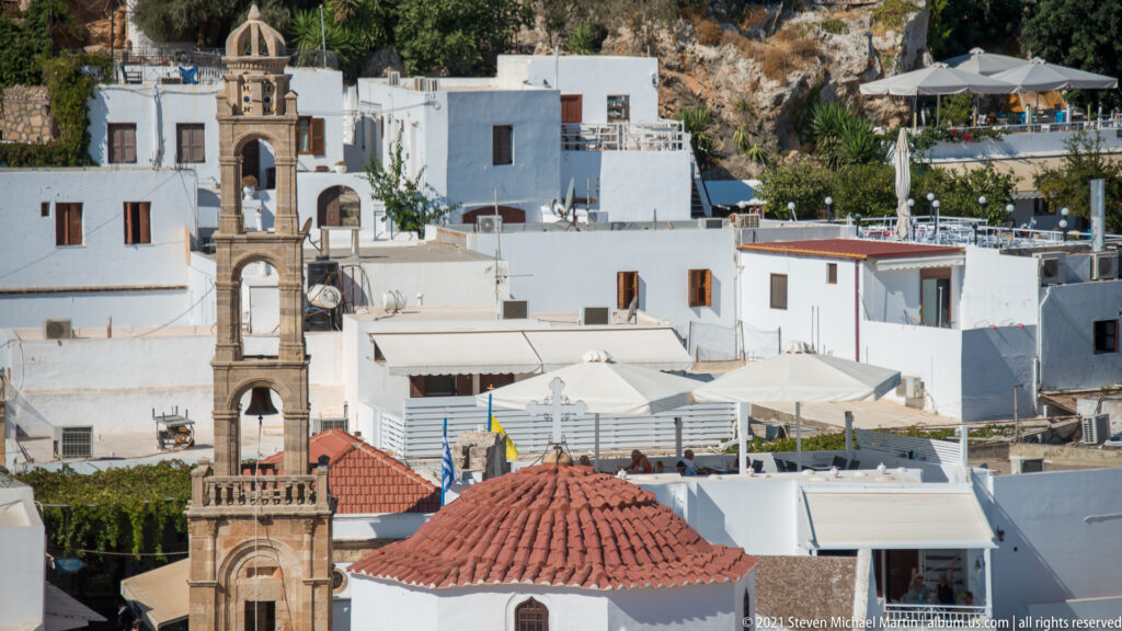 Whitewashed Lindos houses and terraces surrounding stone church bell towers and a red-tiled dome on the hillside.
