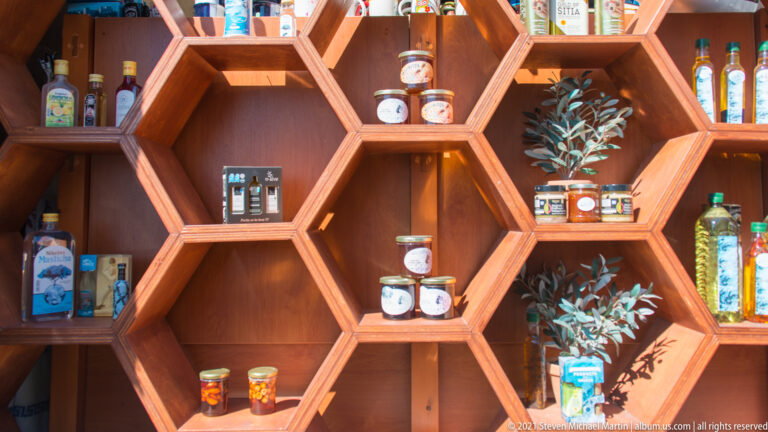 Hexagonal wooden shelves holding jars of preserves or honey, bottles of olive oil and spirits, and decorative olive branches.