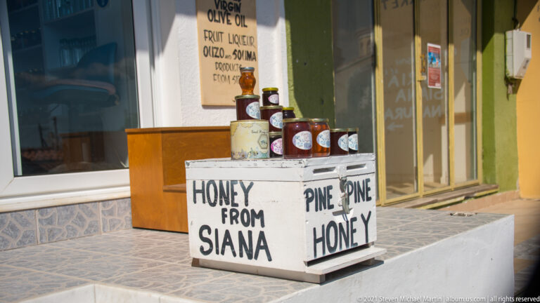 Jars stacked on a white stand marked “HONEY FROM SIANA” and “PINE HONEY” outside a village shop.