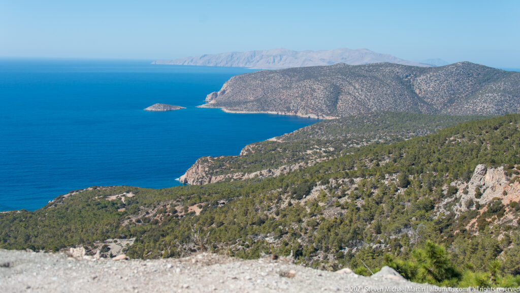 Rugged pine-covered headlands and pale cliffs extend into the blue Aegean, with a small rocky islet offshore.