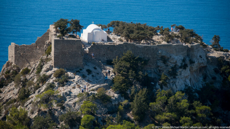 Close view of Monolithos Castle walls on a limestone crag, with a white chapel, trees, steps, and visitors.
