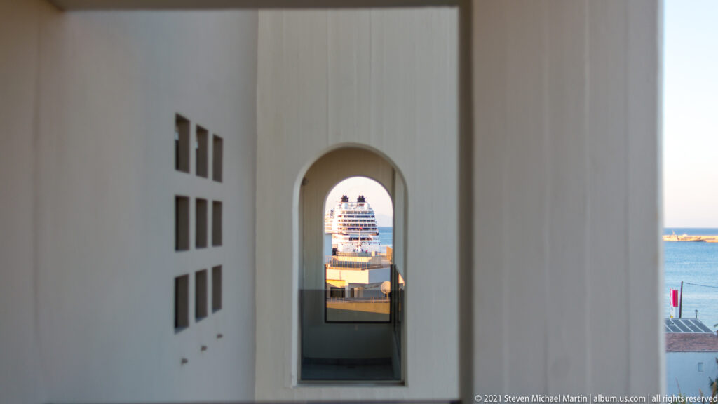 Arched opening in a pale wall framing a cruise ship and blue harbor water beyond.