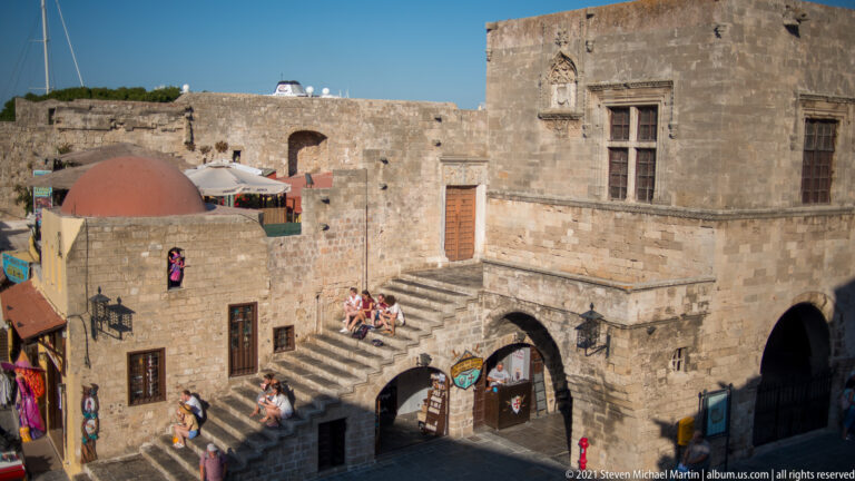 Historic stone building with a steep exterior stair, arched shop openings, seated visitors, and red dome behind.