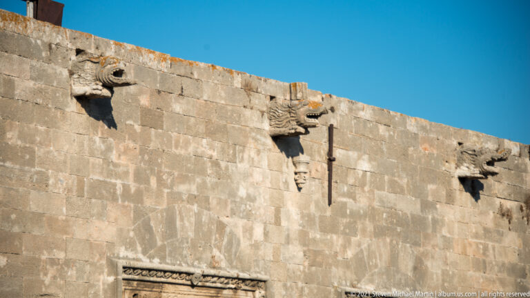 Three carved stone animal-like spouts projecting from the top of a high masonry wall against blue sky.