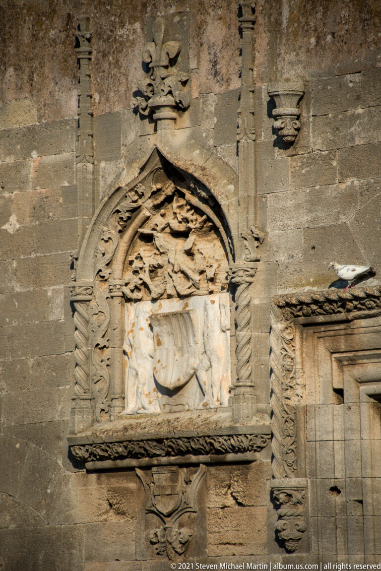 Weathered heraldic shield beneath an ornate pointed leafy canopy and fleur-de-lis-shaped finial on a stone wall.