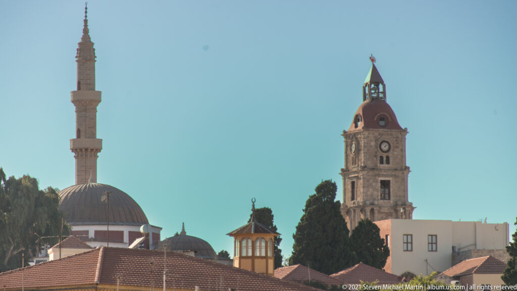 Minaret, domed mosque, and tall clock tower rising above red-tiled roofs and cypress trees.