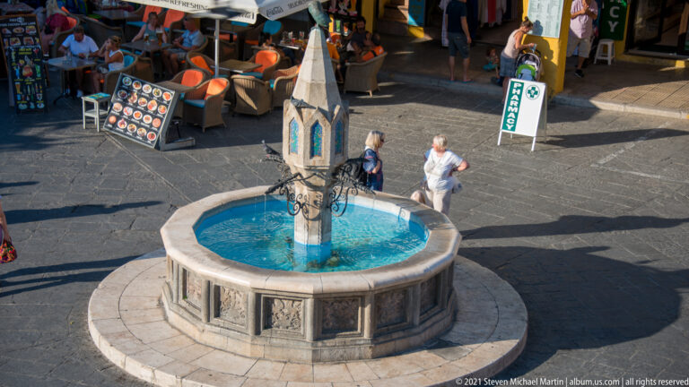 Octagonal stone fountain with blue water, relief panels, and a tall pointed central structure in a public square.