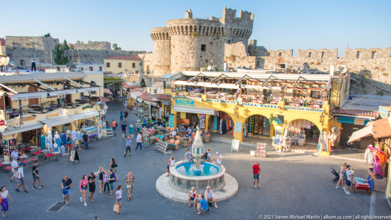 Busy square with central fountain, shops, pedestrians, and a large crenellated round tower behind.