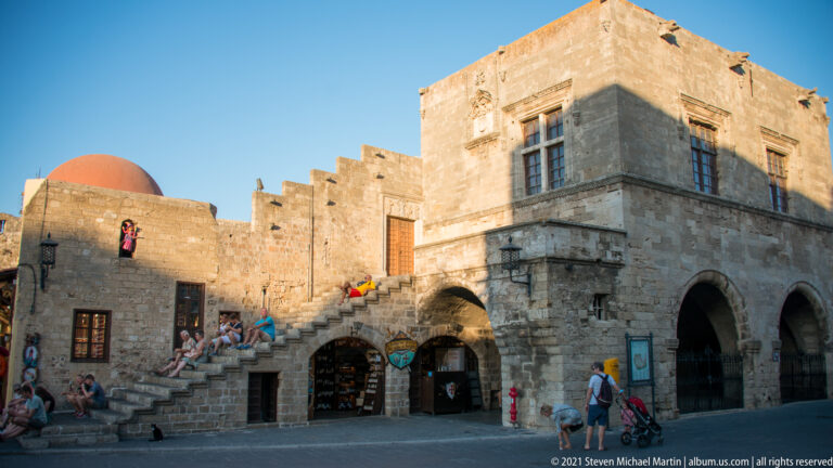 Two-story stone building with exterior stairs, arched lower openings, tall windows, seated visitors, and a red dome.
