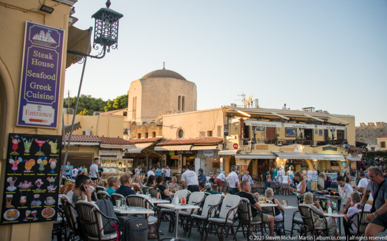 Busy outdoor restaurant seating with diners, menu signs, and a rounded stone historic building behind.
