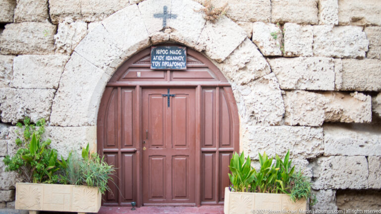 Red pointed wooden chapel door beneath a cross and Greek inscription in rough stonework.