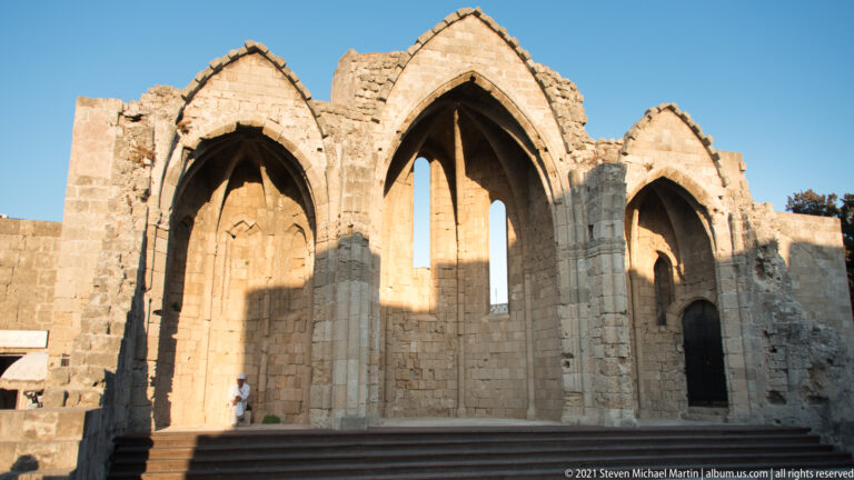 Roofless medieval church ruins with three tall pointed Gothic arches, lancet openings, and broad entrance steps.