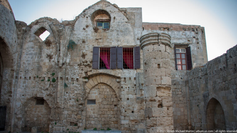 Weathered stone façade with cylindrical projecting tower, shuttered red-curtained window, and blocked arched opening.