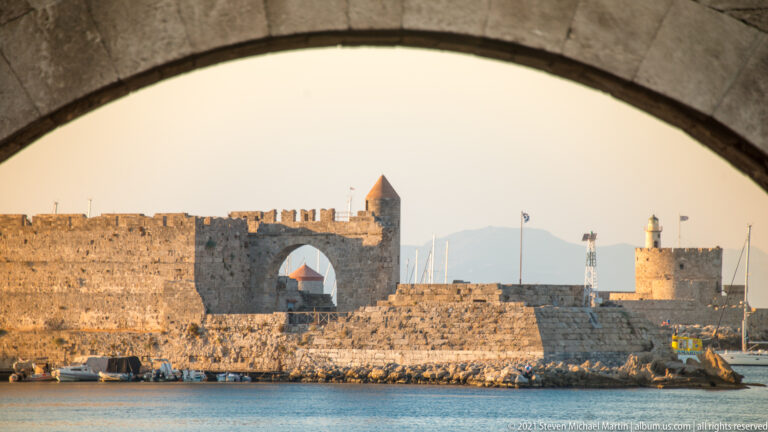 Harbor walls, tower, and lighthouse framed through a broad stone arch above blue water.