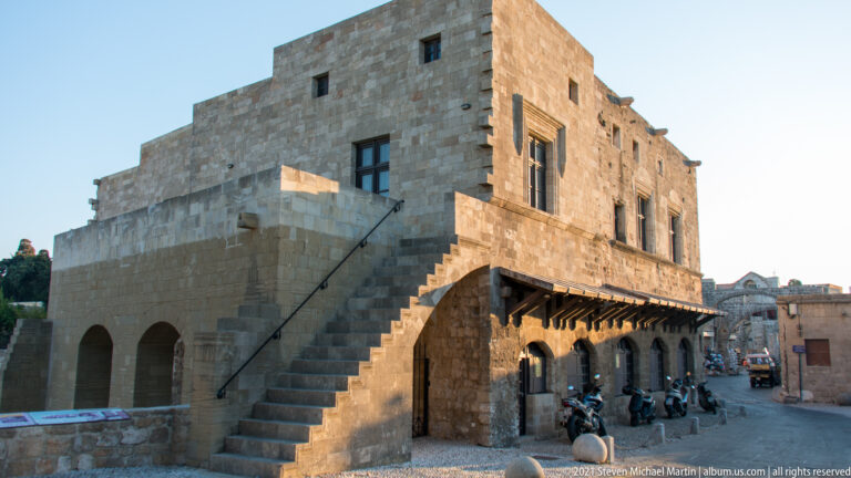 Large stone building with long exterior staircase, arched lower level, timber overhangs, and scooters below.