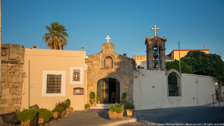 Whitewashed Orthodox chapel entrance with crosses, painted icon, bell frame, potted plants, and palm tree.