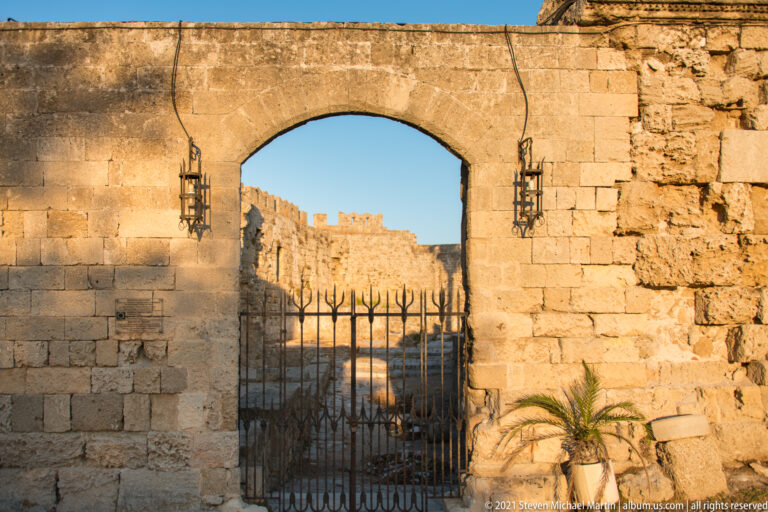 Broad stone arch with iron gate and wall lanterns, framing sunlit battlements beyond.