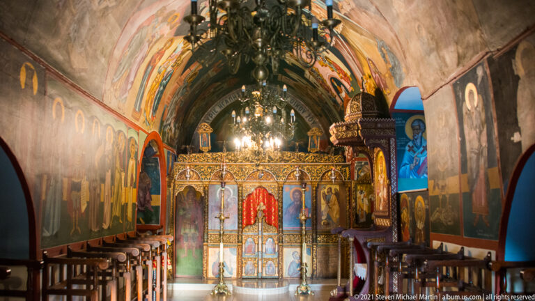 Orthodox church interior with gilded iconostasis, hanging chandelier, painted saints, and colorful vaulted ceiling.