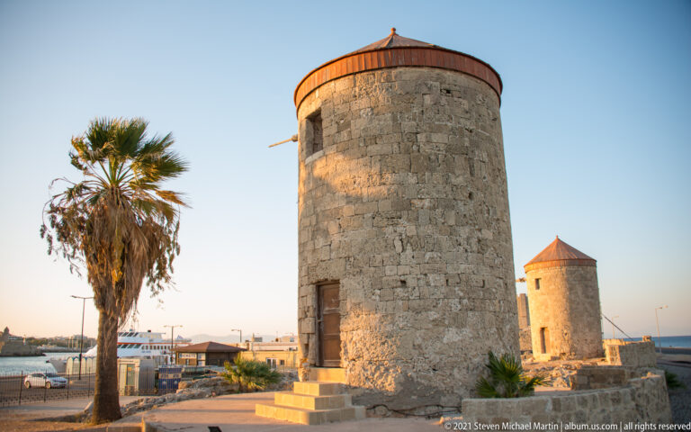 Two cylindrical stone windmills with conical roofs along the harbor, with a tall palm beside the nearer mill.