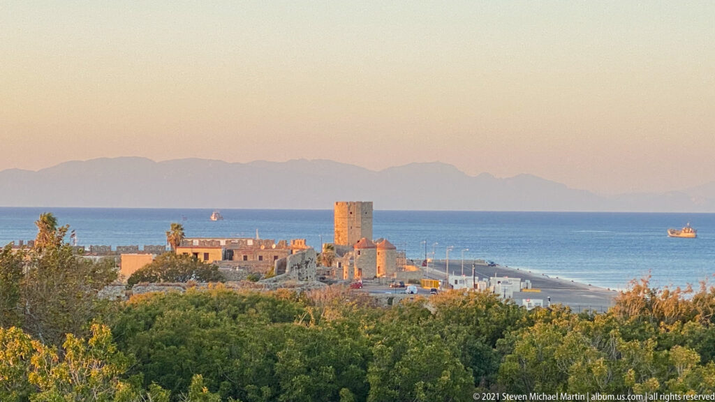 Elevated coastal view over treetops toward harbor buildings, blue sea, offshore vessel, and hazy distant mountains.
