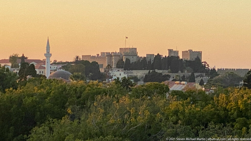 Rhodes Old Town skyline at sunset with minaret, mosque dome, crenellated walls, towers, and trees.