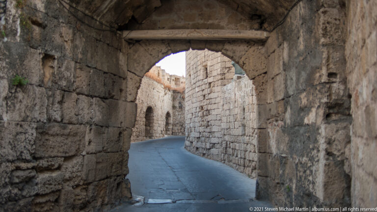 View from inside a dark stone archway onto a narrow curving lane between high walls.