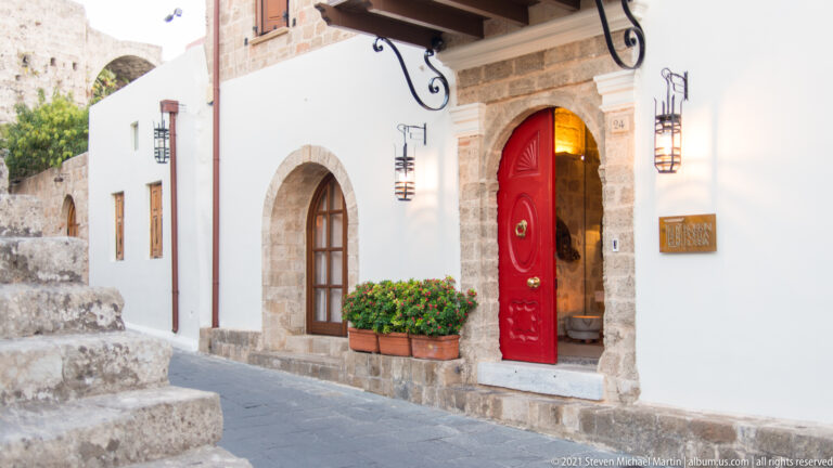Whitewashed façade with stone-trimmed arches, a bright red door, lanterns, and potted plants.