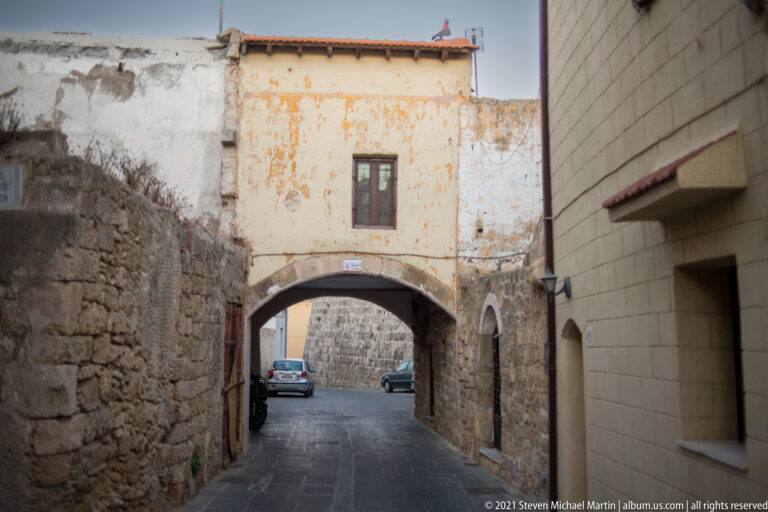 Weathered building bridging a narrow lane on a broad arch, with parked cars beyond.