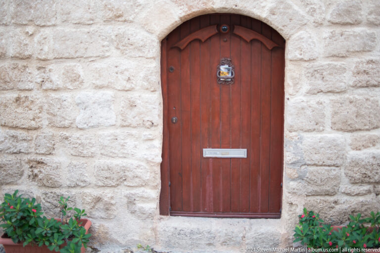 Red-brown arched wooden door with a mail slot and knocker in a pale stone wall.
