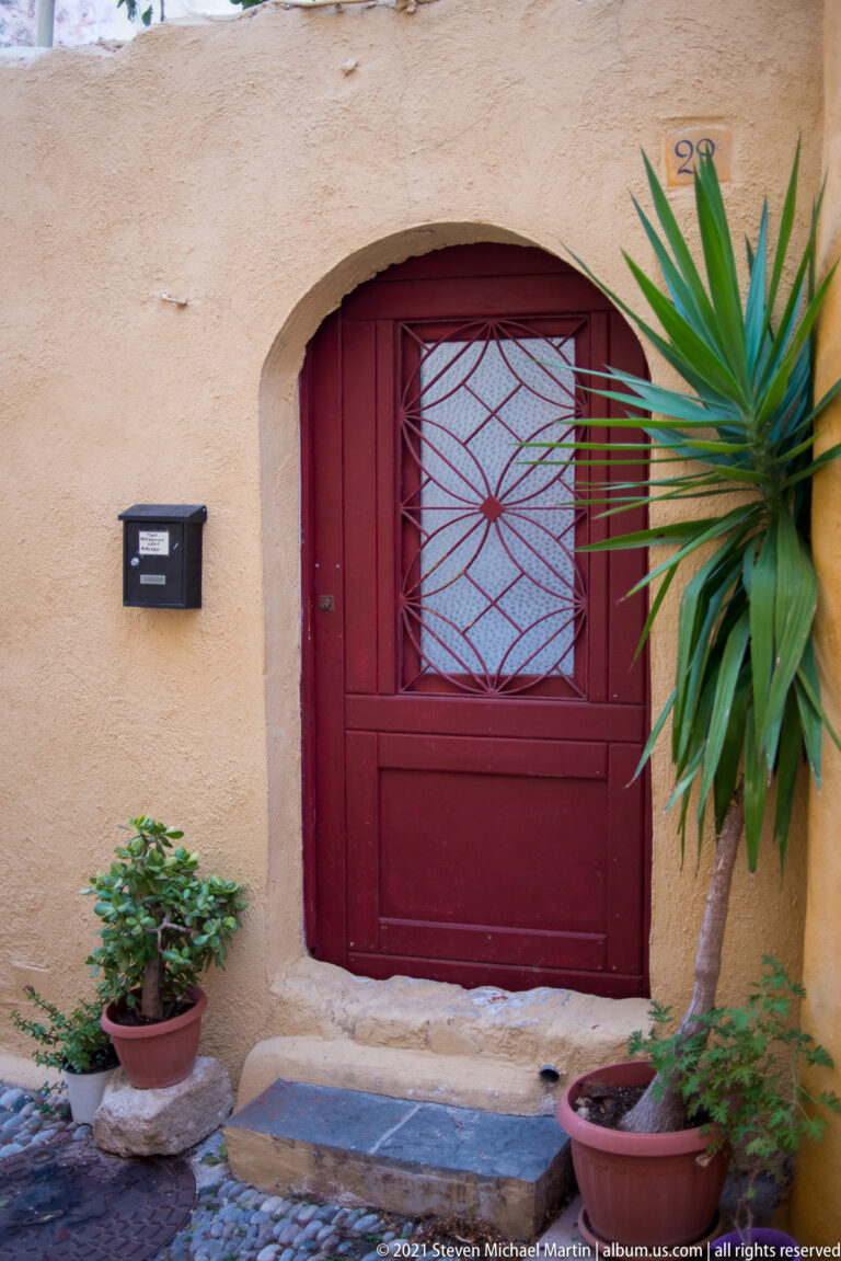 Red recessed door with a geometric metal grille, mailbox, and potted plants.