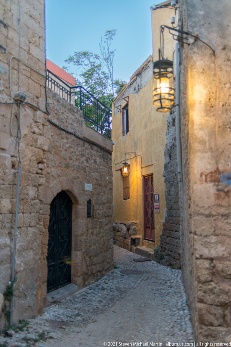 Narrow cobbled lane between stone buildings, lit by warm wall lanterns at dusk.